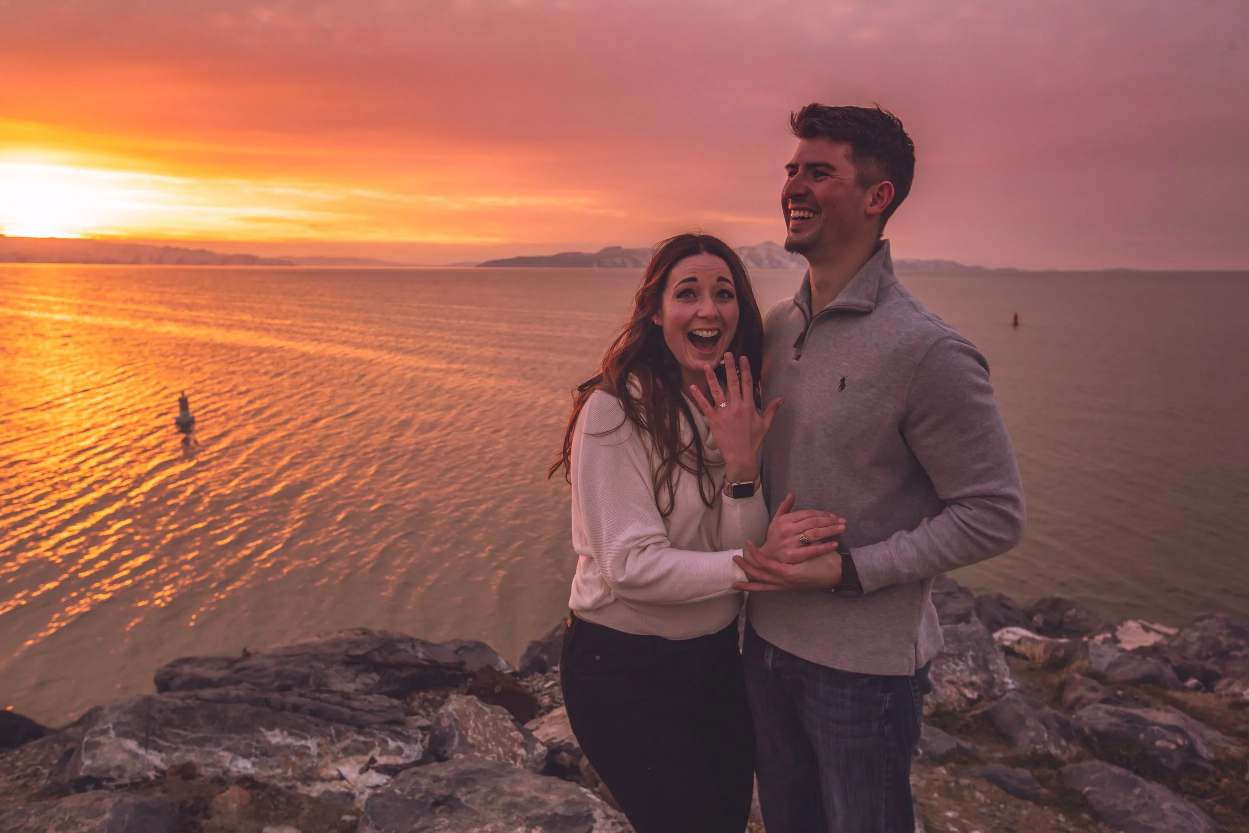 A couple standing on rocks by the water during sunset, with the woman showing a ring on her finger and both smiling happily.