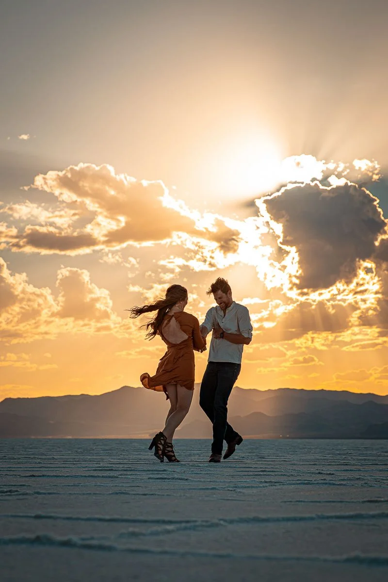 A couple dancing on a flat surface during sunset, with mountains in the background and clouds in the sky.