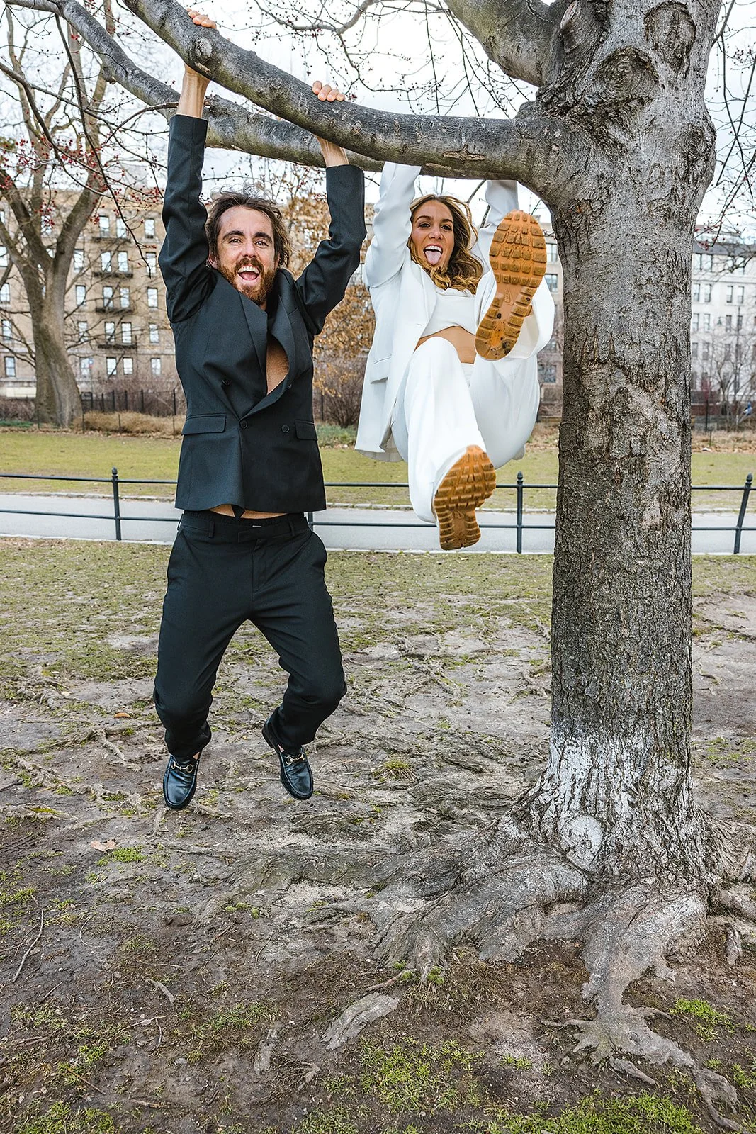 A man in a black suit and a woman in a white suit hanging from a tree branch in a park, smiling and making playful faces.