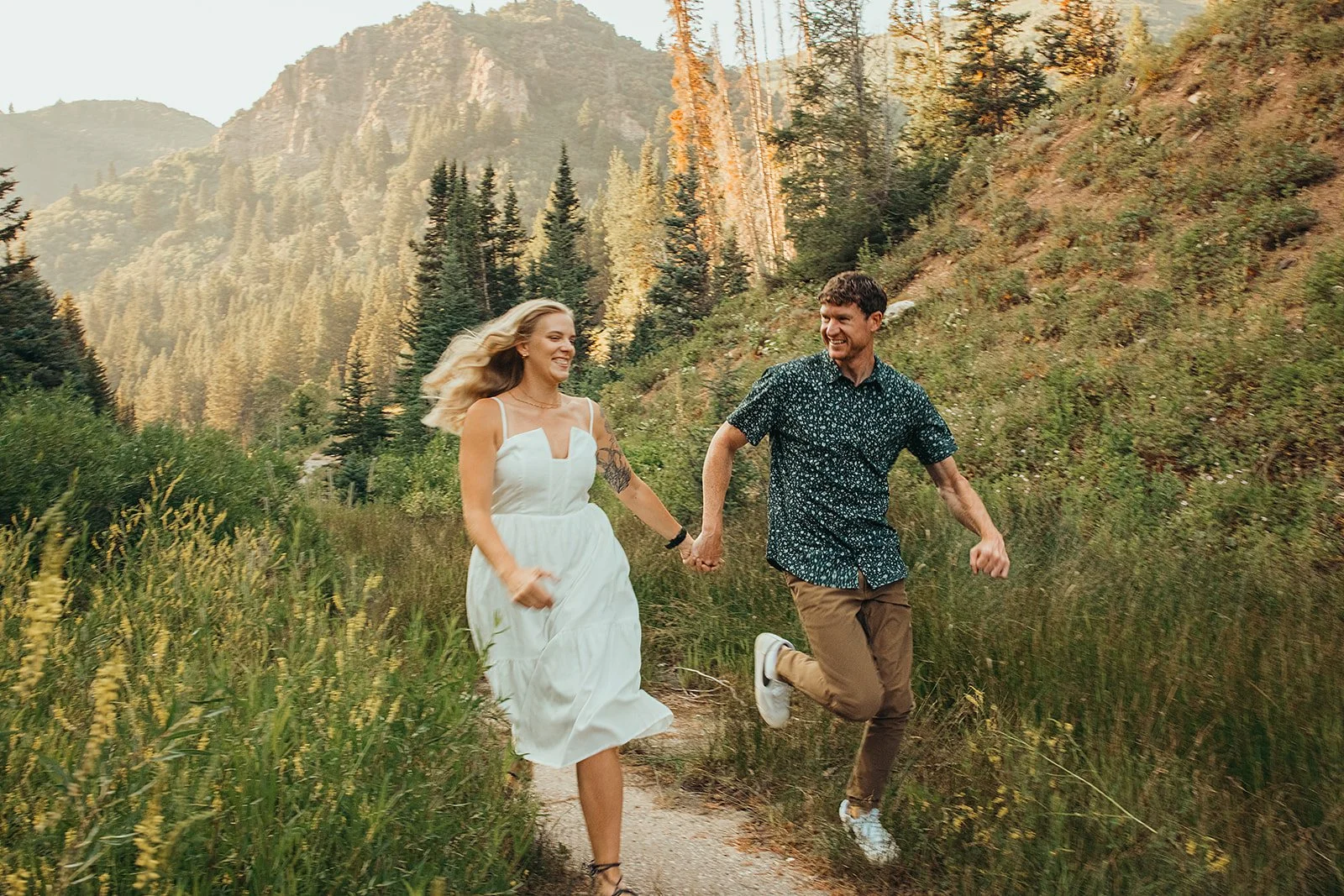 A happy couple running hand in hand on a nature trail surrounded by greenery and mountains during sunset.