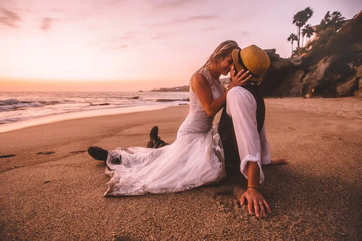 A couple sits on the beach at sunset, with the woman wearing a white dress and the man wearing a vest, a hat, and rolled-up sleeves. They are touching foreheads and smiling.