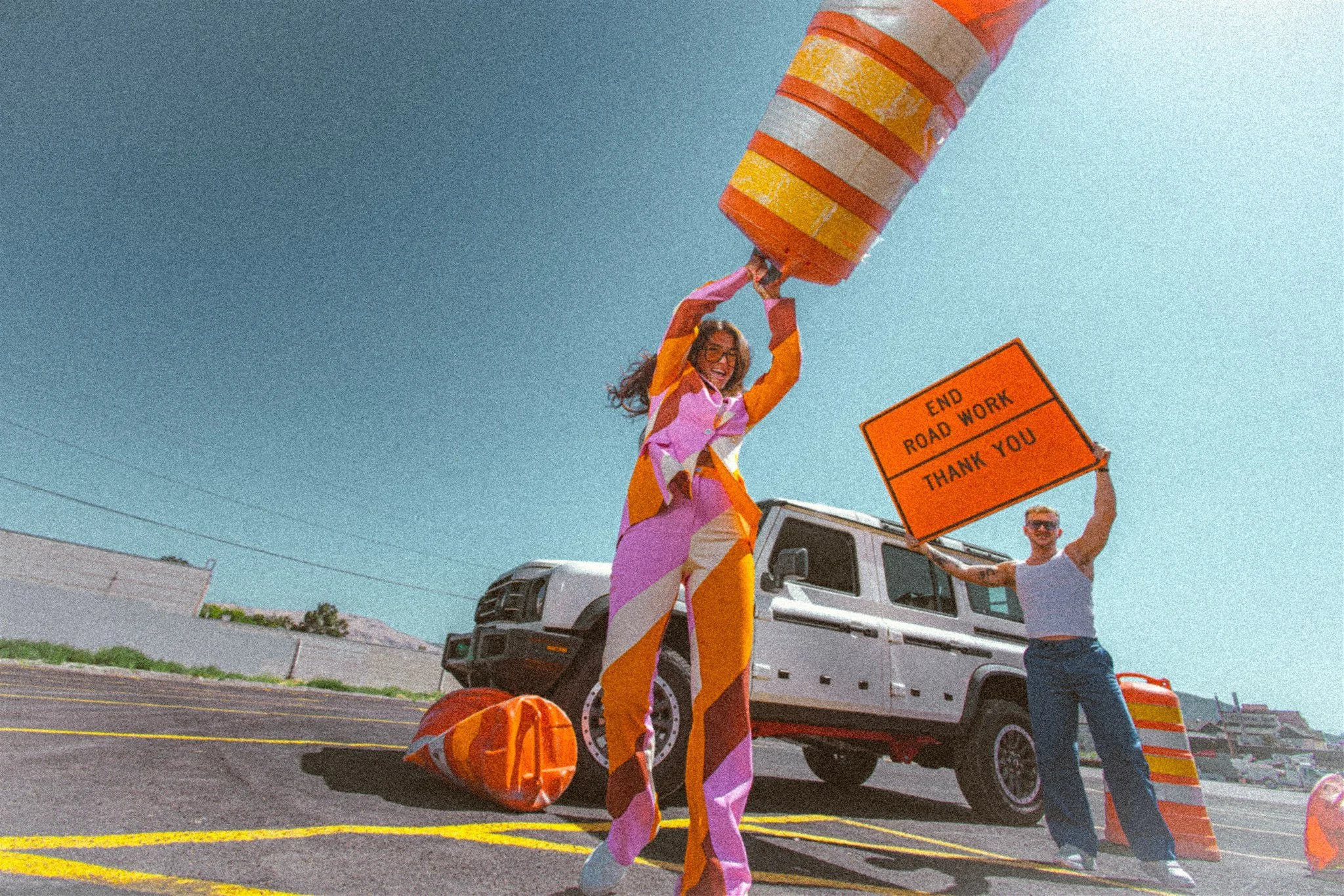 Two people celebrating at a roadwork site with orange traffic barrels, one holding a sign that says 'End Road Work Thank You,' and the other raising a traffic barrel above her head, with a white truck in the background and a clear blue sky.