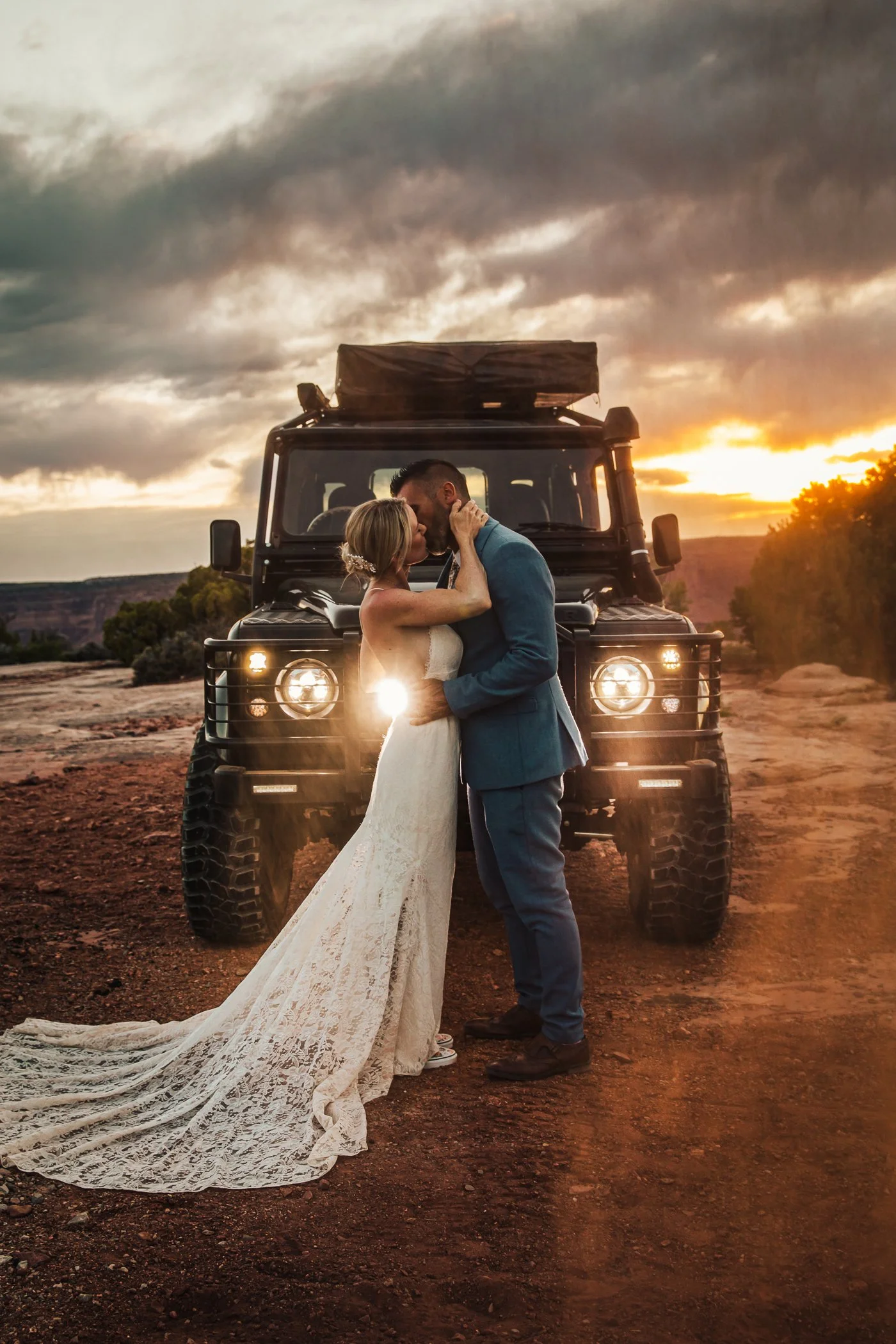 A bride and groom kissing in front of a black off-road vehicle during sunset in a desert landscape.