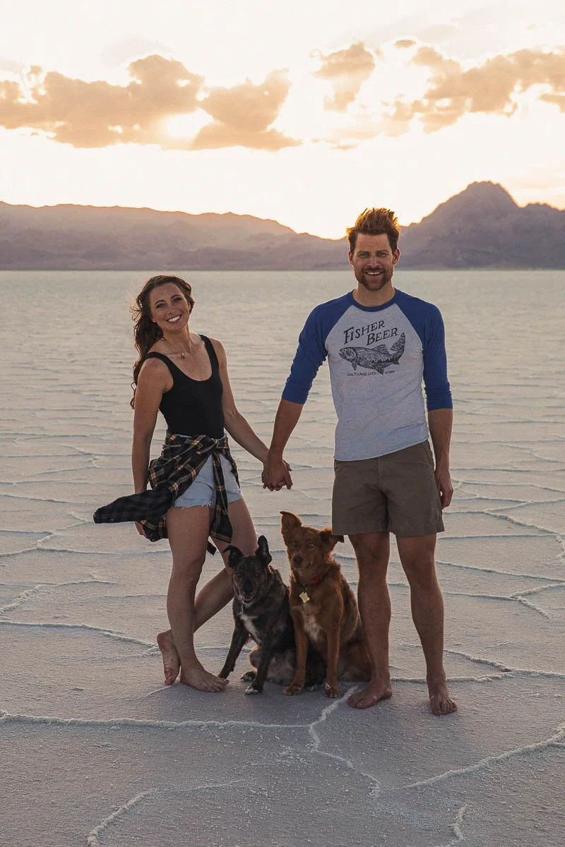 A smiling couple holding hands on a salt flat at sunset, accompanied by two dogs, with mountains in the background.