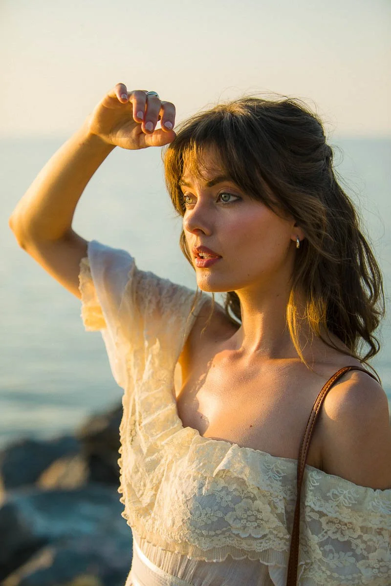 A woman with brown hair and light skin standing outdoors near water, looking into the distance, with her right arm raised and her hand near her forehead, bathed in warm sunlight.