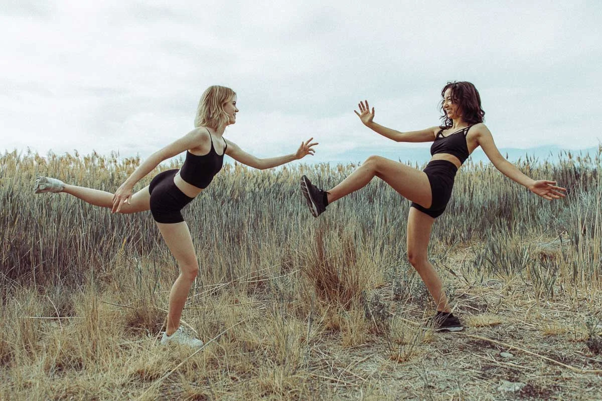 Two women in black athletic wear are playing in a grassy field, with one woman holding the other's ankle while she balances on one leg.