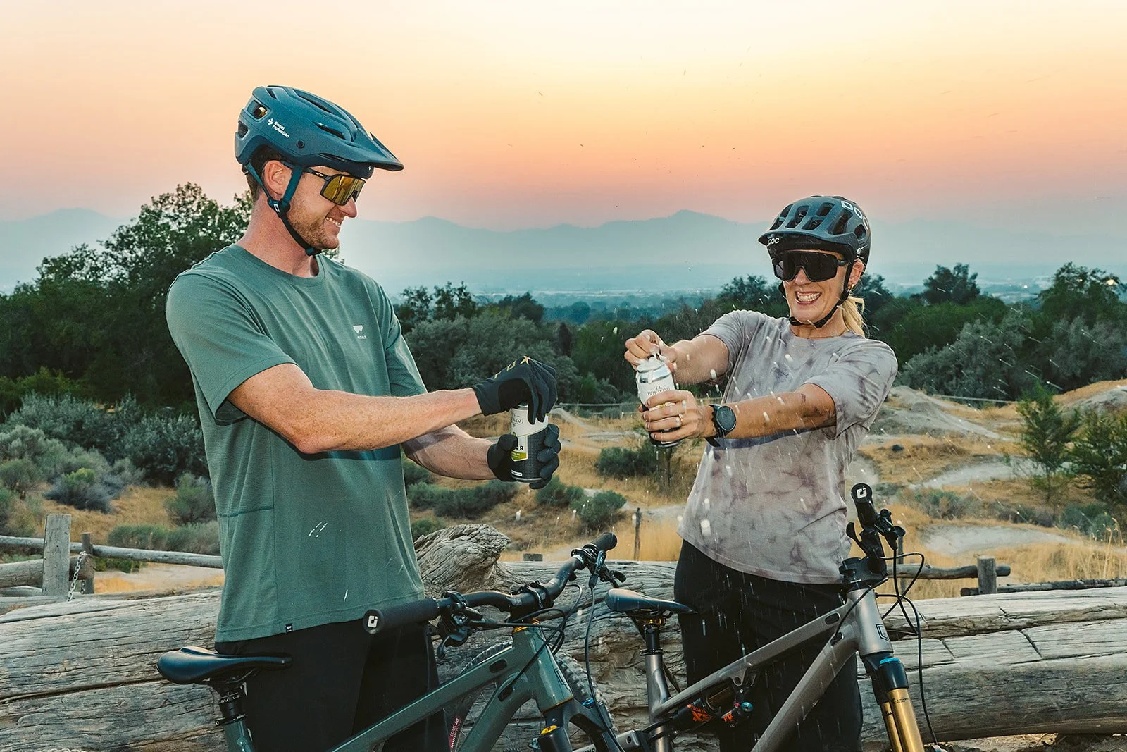 A man and woman in cycling gear, wearing helmets and sunglasses, are outdoors on mountain bikes. The woman is opening a bottle, spraying water, while the man is holding another bottle. They are standing near a wooden fence with scenic hills and trees