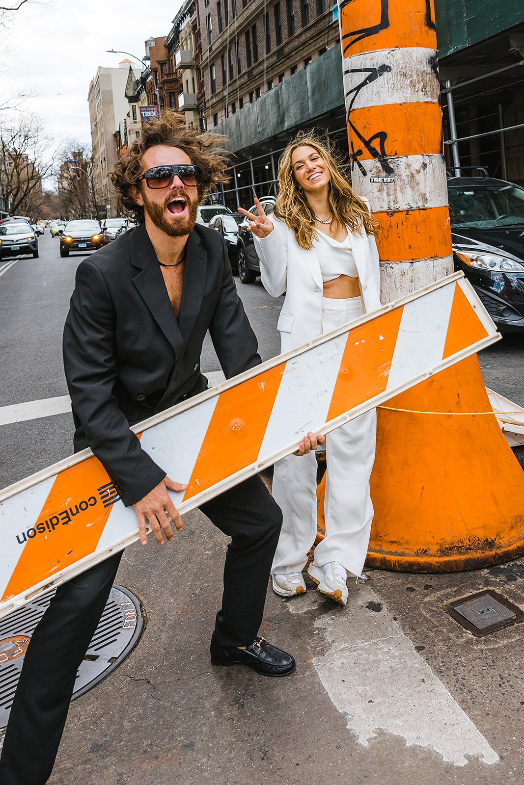 A man in a black suit and sunglasses holding a safety barrier, next to a woman in a white suit making a peace sign, standing on a city street with cars and buildings in the background.
