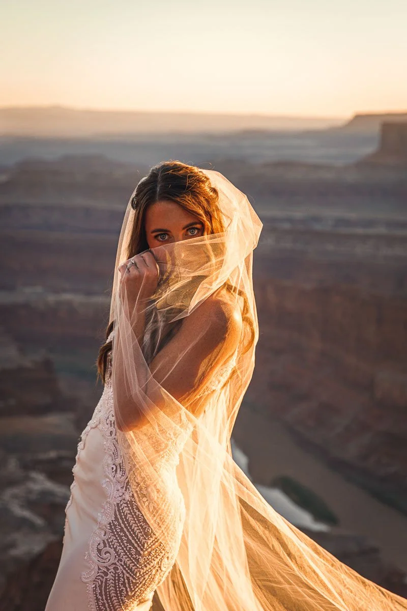 A woman with long brown hair and blue eyes, wearing a white lace dress, is partially covered with a sheer, golden-orange fabric. She stands in a desert landscape at sunset, with the setting sun casting a warm glow on her and the canyon behind her.