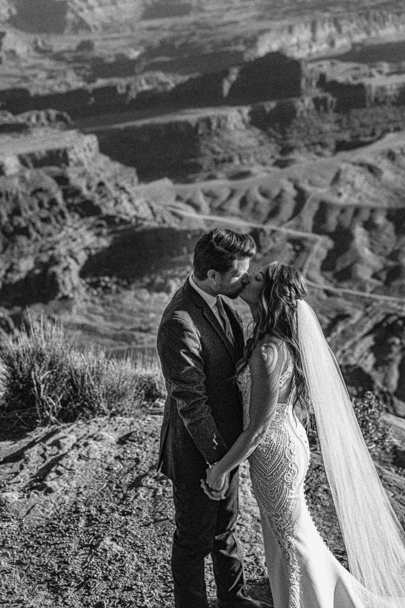 A black-and-white photo of a bride and groom sharing a kiss on a hilltop at the Grand Canyon, holding hands with the canyon visible in the background.