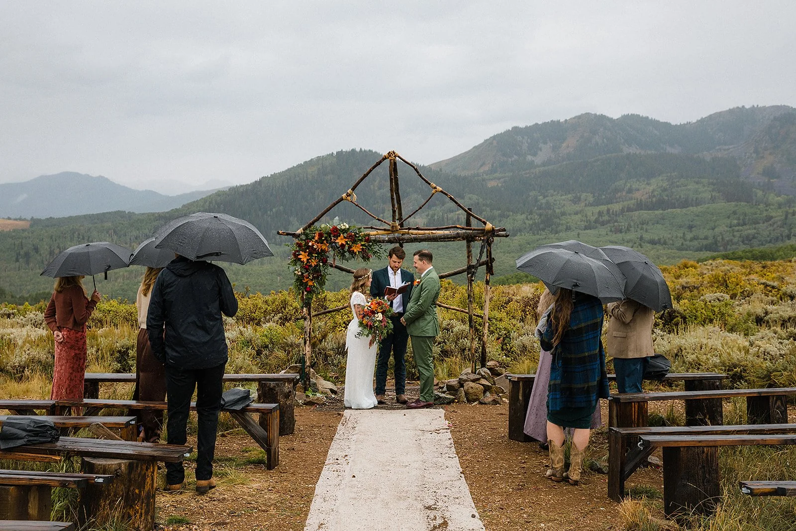 A wedding ceremony outdoors on a rainy day with guests holding umbrellas, a couple standing under a decorated wooden arch, and a scenic mountain landscape in the background.
