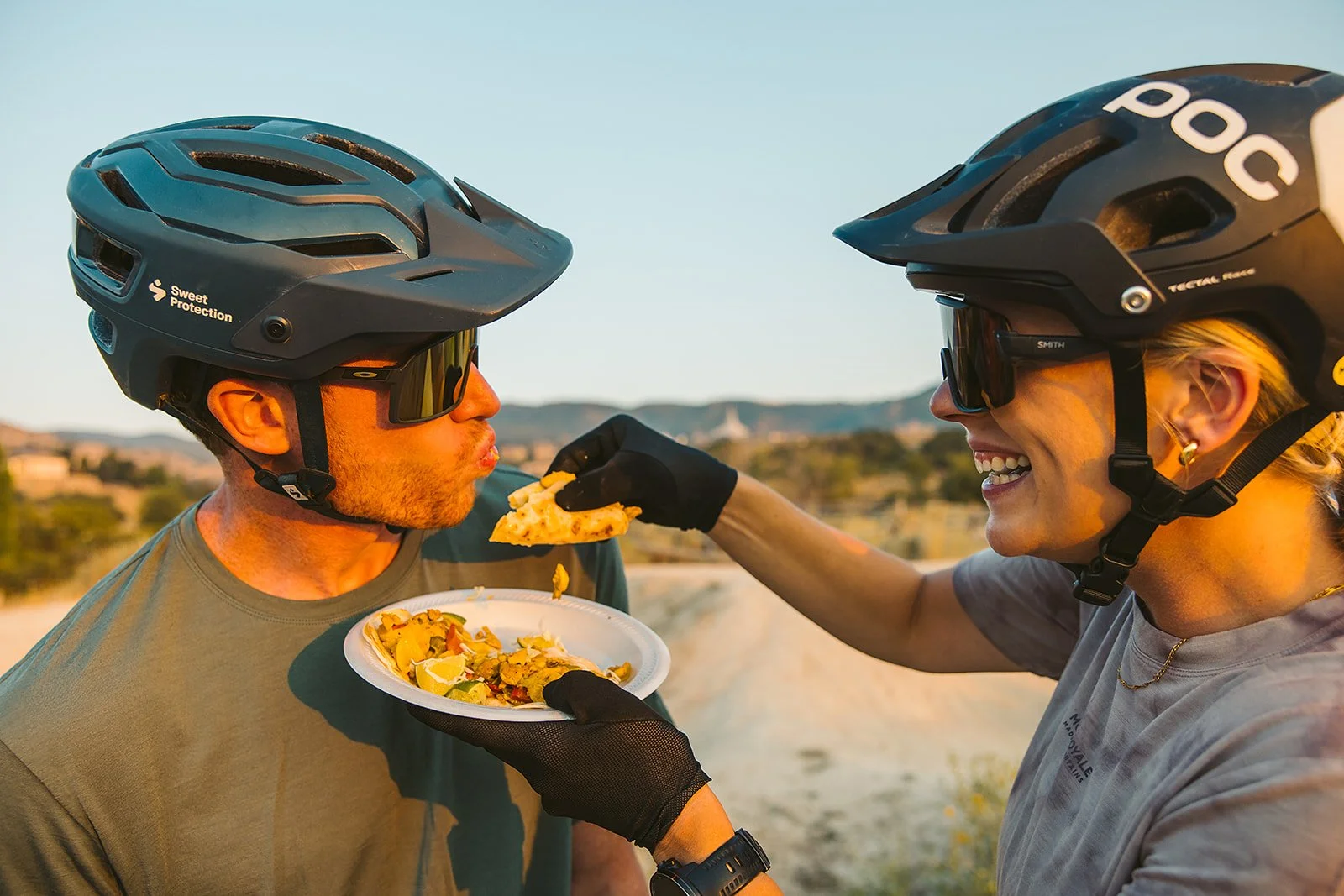Two cyclists wearing helmets and sunglasses sharing a slice of pizza outdoors, with a plate of food and a bright sky in the background.
