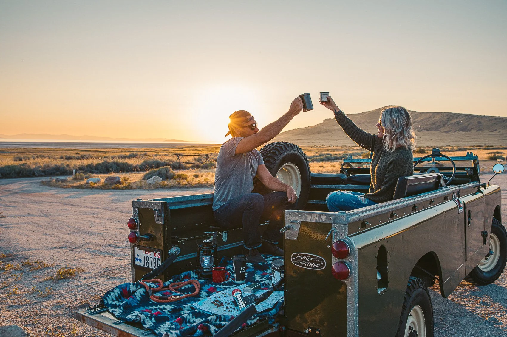 A man and woman sitting in the back of a truck, toasting each other with coffee mugs during sunset in a desert landscape.