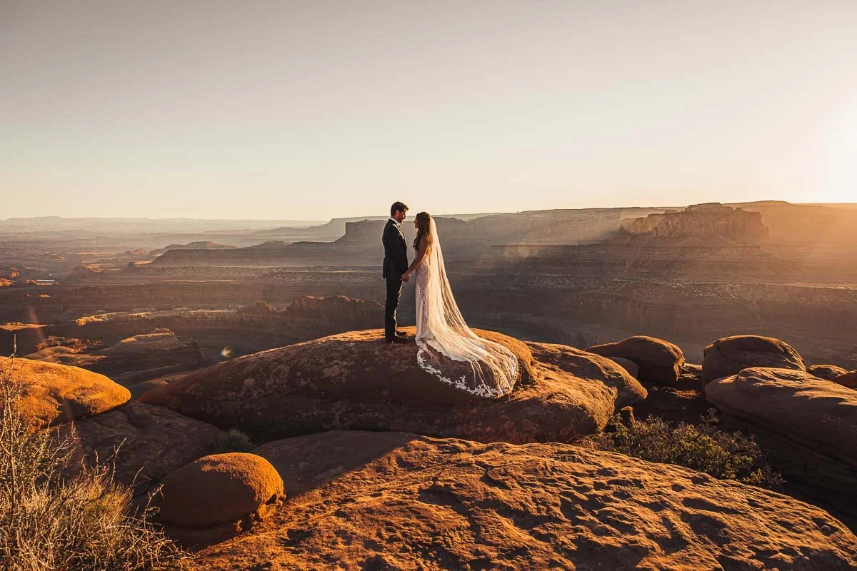 A bride and groom holding hands on a large rock at sunset in front of a canyon landscape.