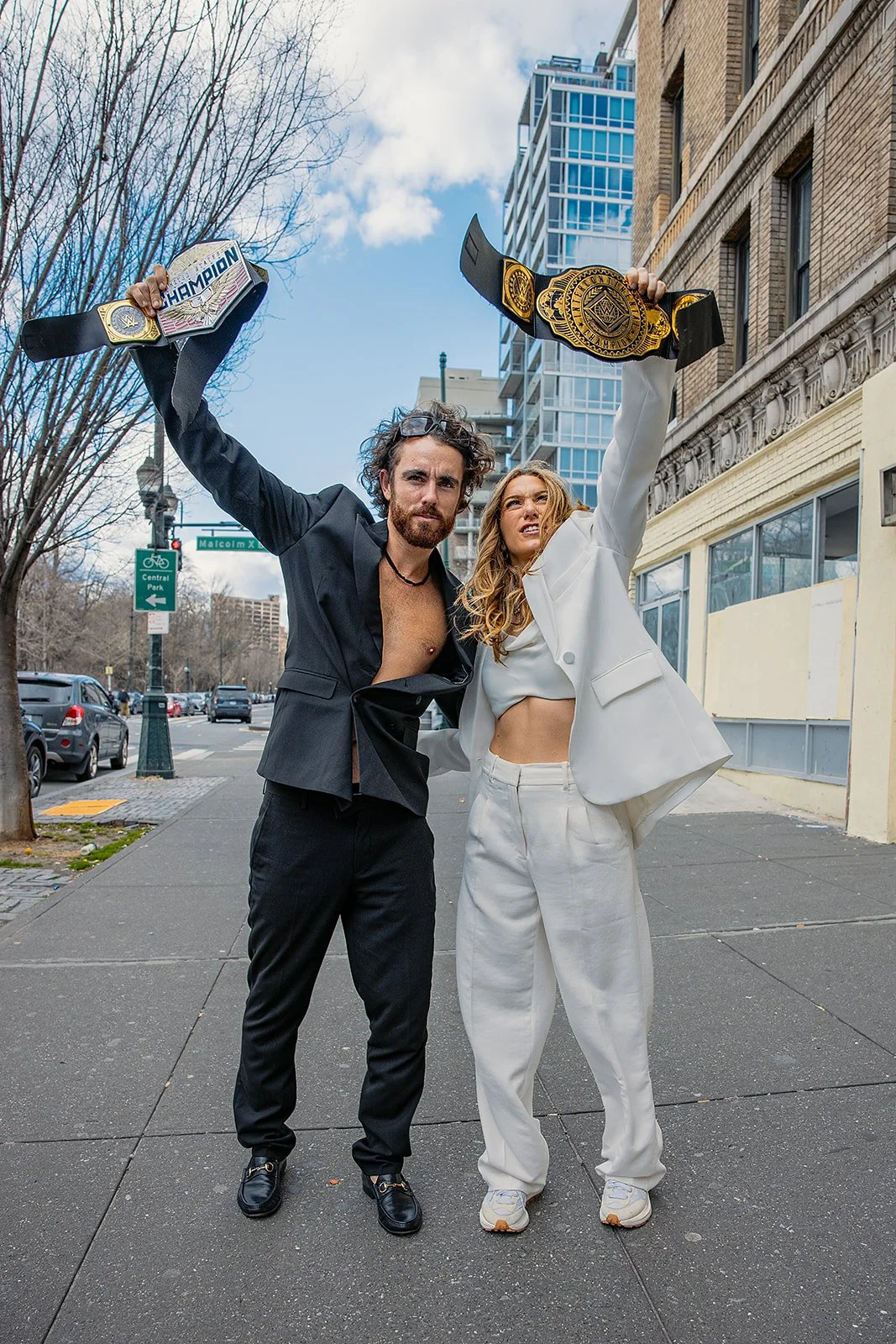 Two people standing on a city sidewalk holding championship belts above their heads, celebrating their victory.