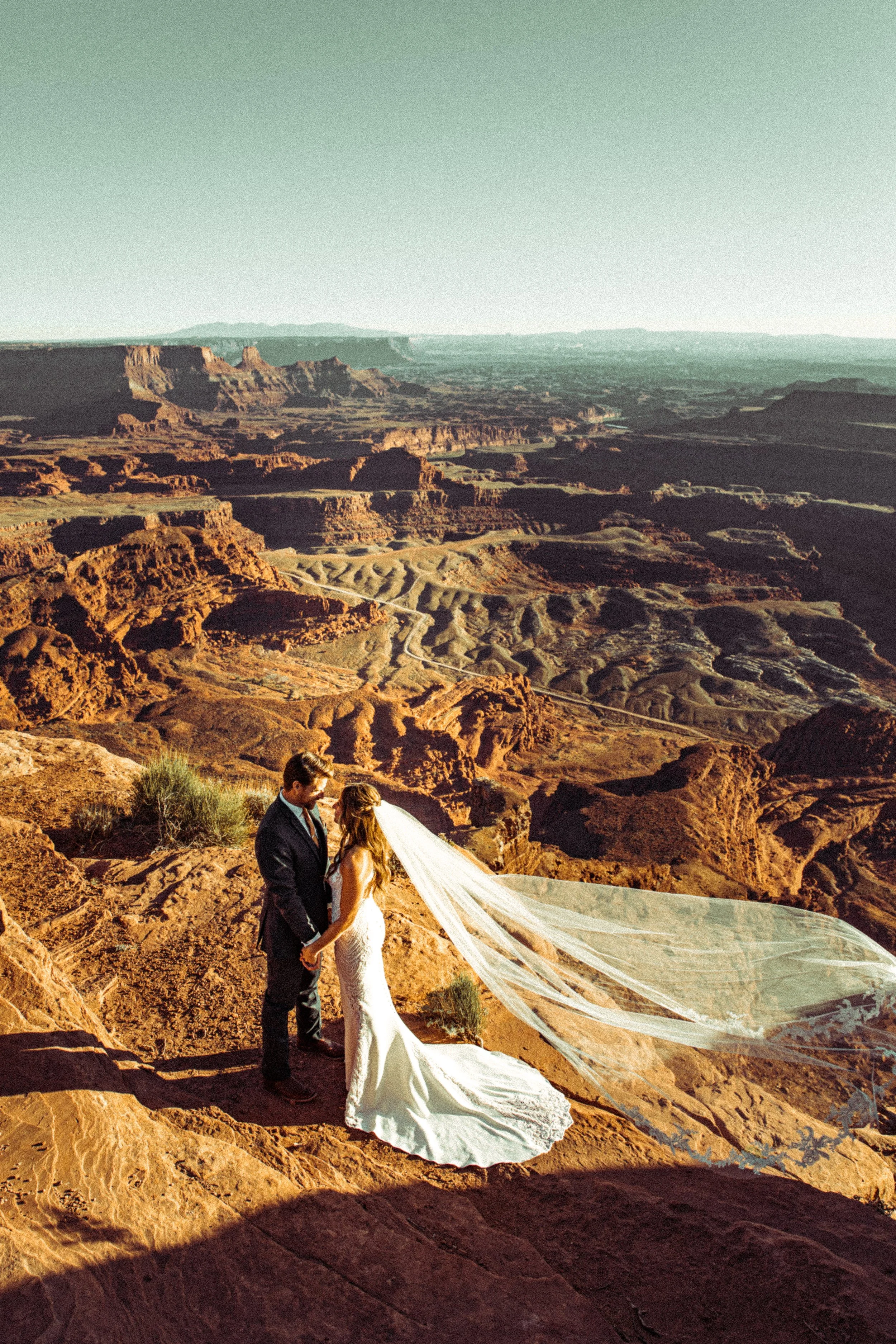A bride and groom holding hands on a rocky ledge in a desert landscape with red rock formations and canyons, during their wedding ceremony.