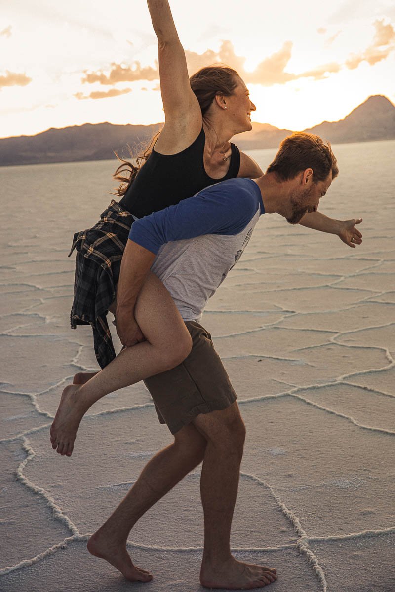 A man giving a woman a piggyback ride on a salt flat during sunset.