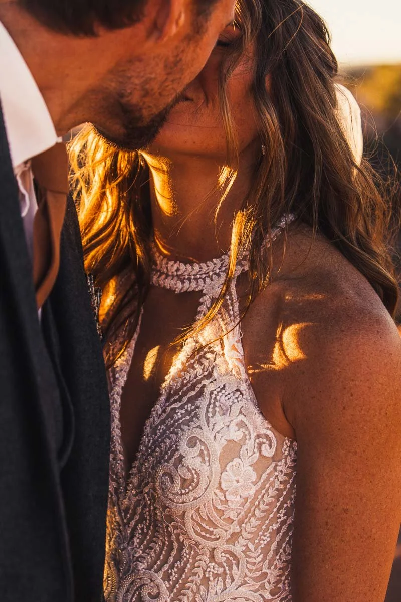 Close-up of a couple kissing, with warm golden lighting highlighting their faces and details of the woman's white lace dress.