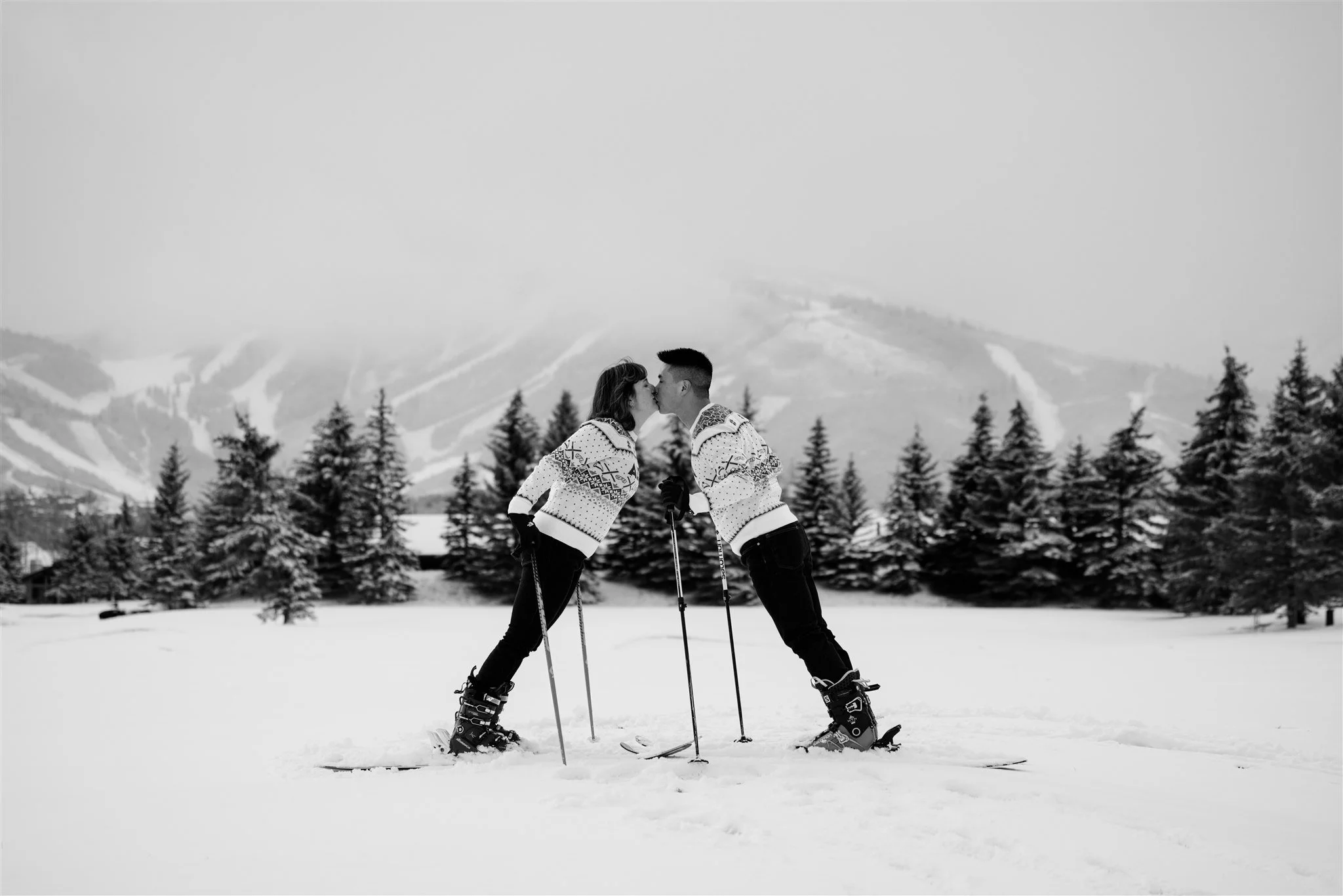 A black-and-white photo of two people on skis in a snowy landscape, leaning in for a kiss, with trees and mountains in the background.