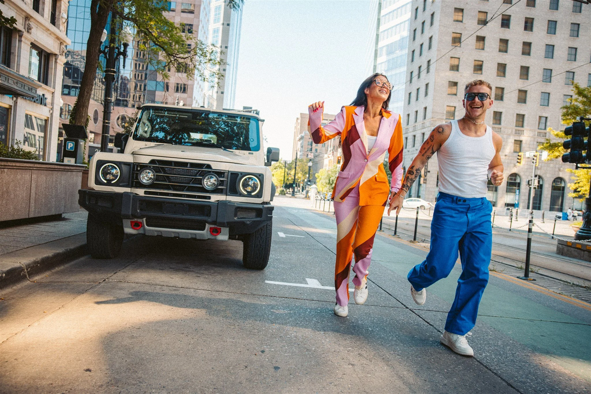 Two cheerful young adults, a woman in a colorful blazer and pants and a man in a white tank top and blue pants, are running and laughing on a city street in daylight, with tall buildings and a parked off-road vehicle behind them.
