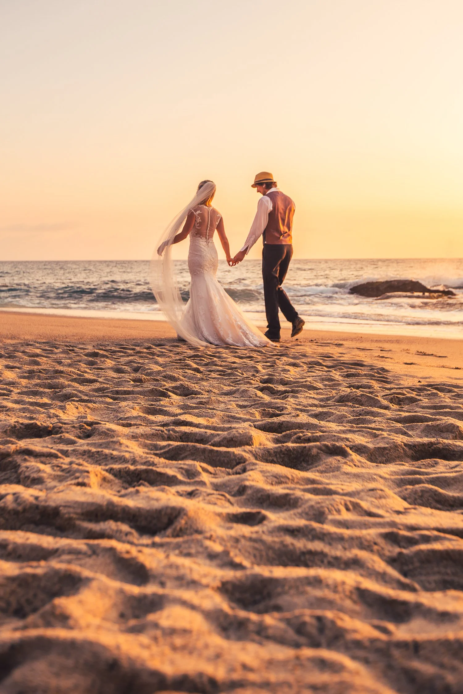 A bride and groom holding hands on the beach at sunset