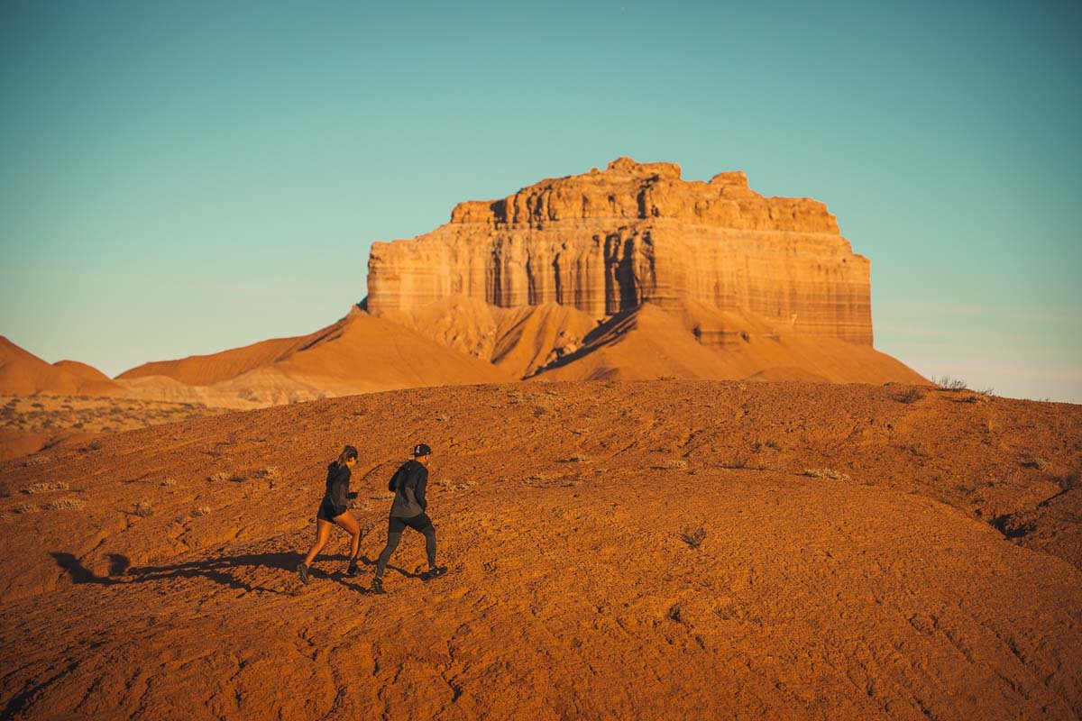 Two hikers walking on a desert trail towards a large rock formation at sunset.