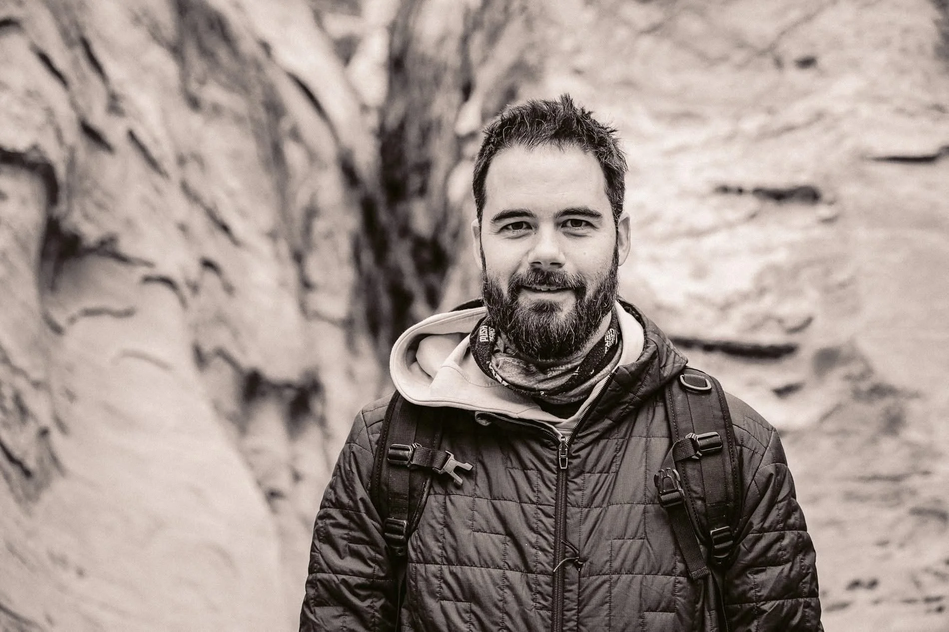 A man with a beard and short hair, wearing a jacket and backpack, smiling outdoors in front of rock formations.