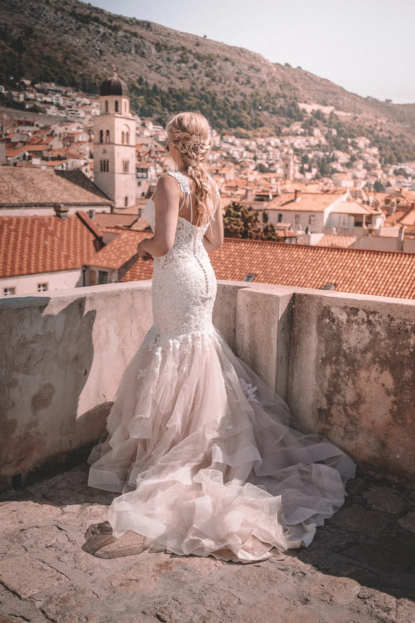 A woman in a wedding dress standing on a rooftop terrace, overlooking a European town with red-tiled roofs and a church with a bell tower, surrounded by hills.