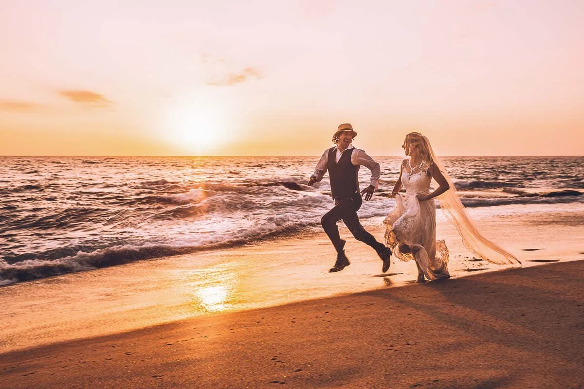 A couple in wedding attire running on the beach at sunset, with the ocean waves behind them.