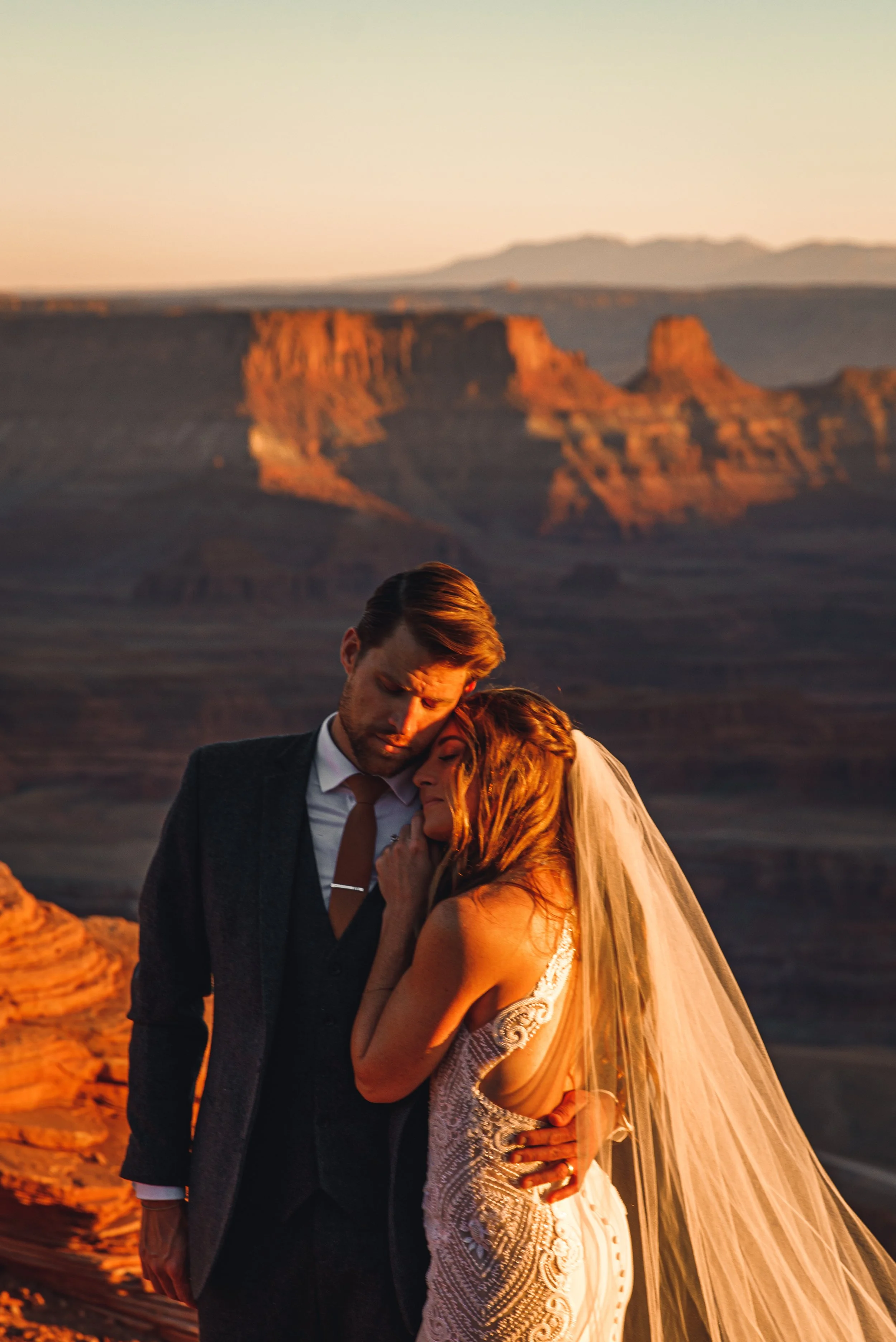A couple dressed in wedding attire embracing at the Grand Canyon during sunset.