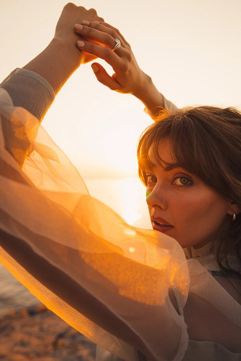 A woman with brown hair and blue eyes posing outdoors during sunset, holding her hands together above her head, with sunlight illuminating her face and sheer orange fabric.