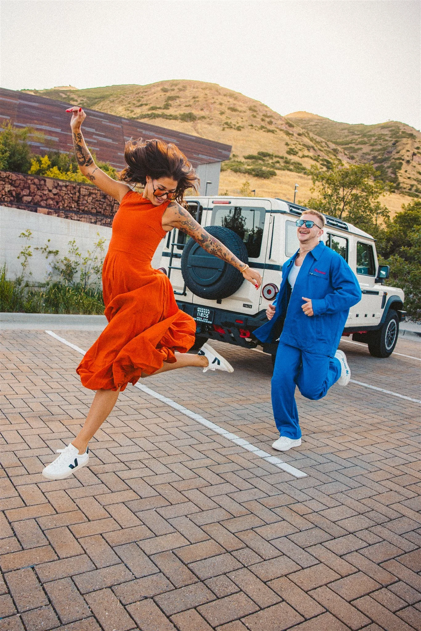 A woman in an orange dress and white sneakers jumping and dancing next to a man in a blue suit who is running beside her in a parking lot with a white SUV and a scenic hillside in the background.