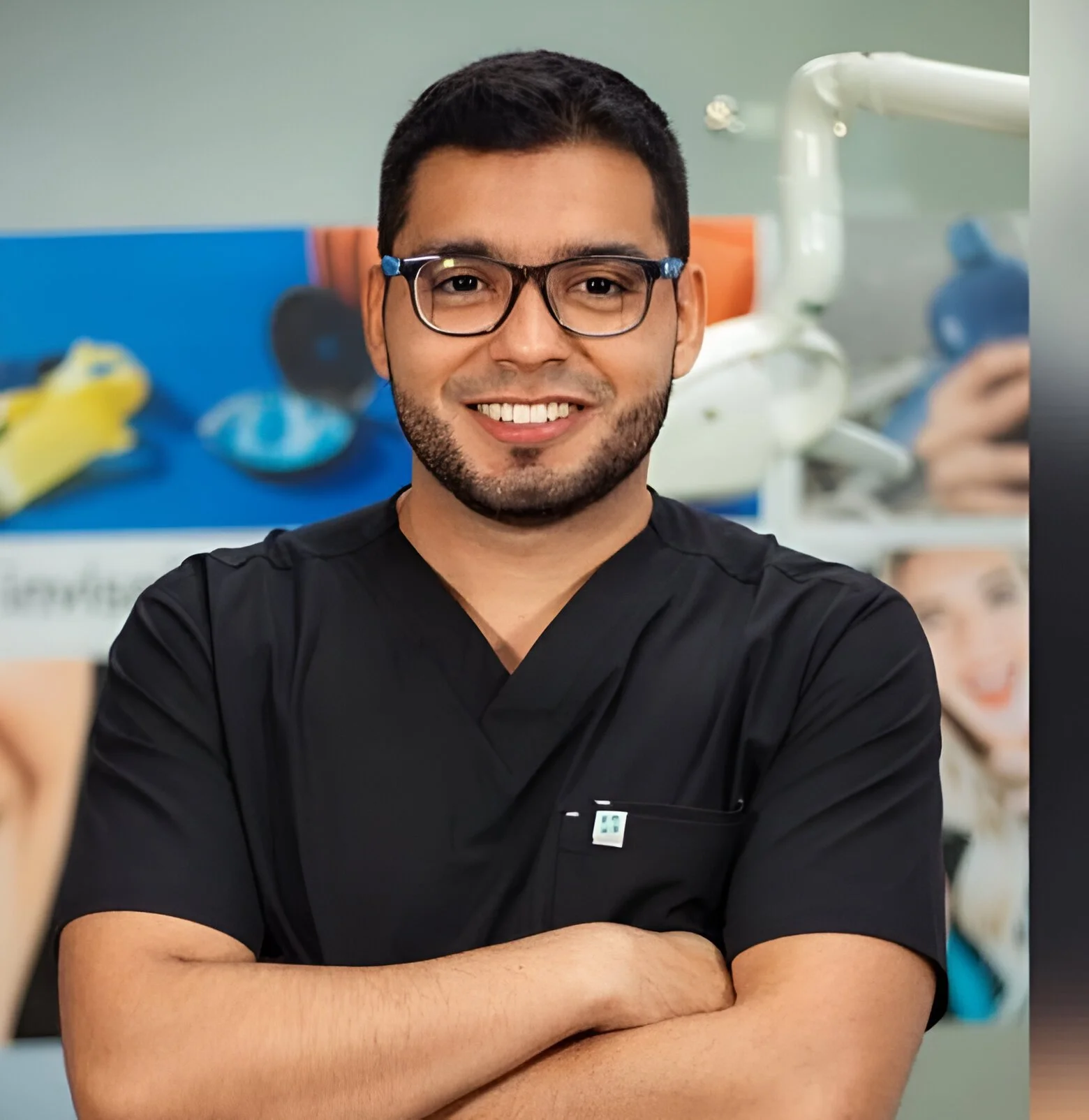 A smiling man wearing glasses and a black medical scrub top standing with arms crossed in a medical or dental office with posters and equipment in the background.