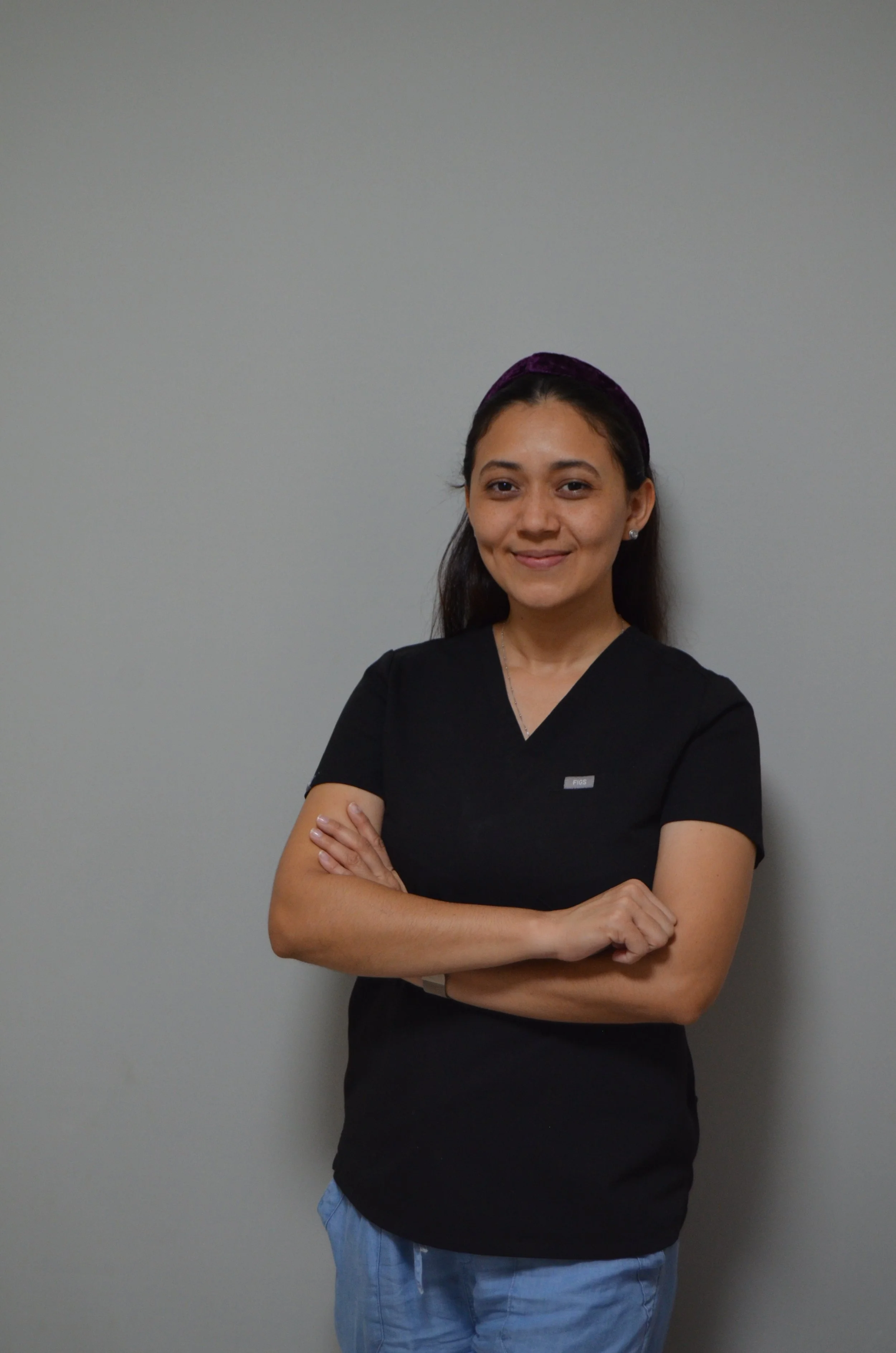 A young woman with dark hair pulled back wearing black medical scrubs, standing against a plain gray wall, smiling with her arms crossed.