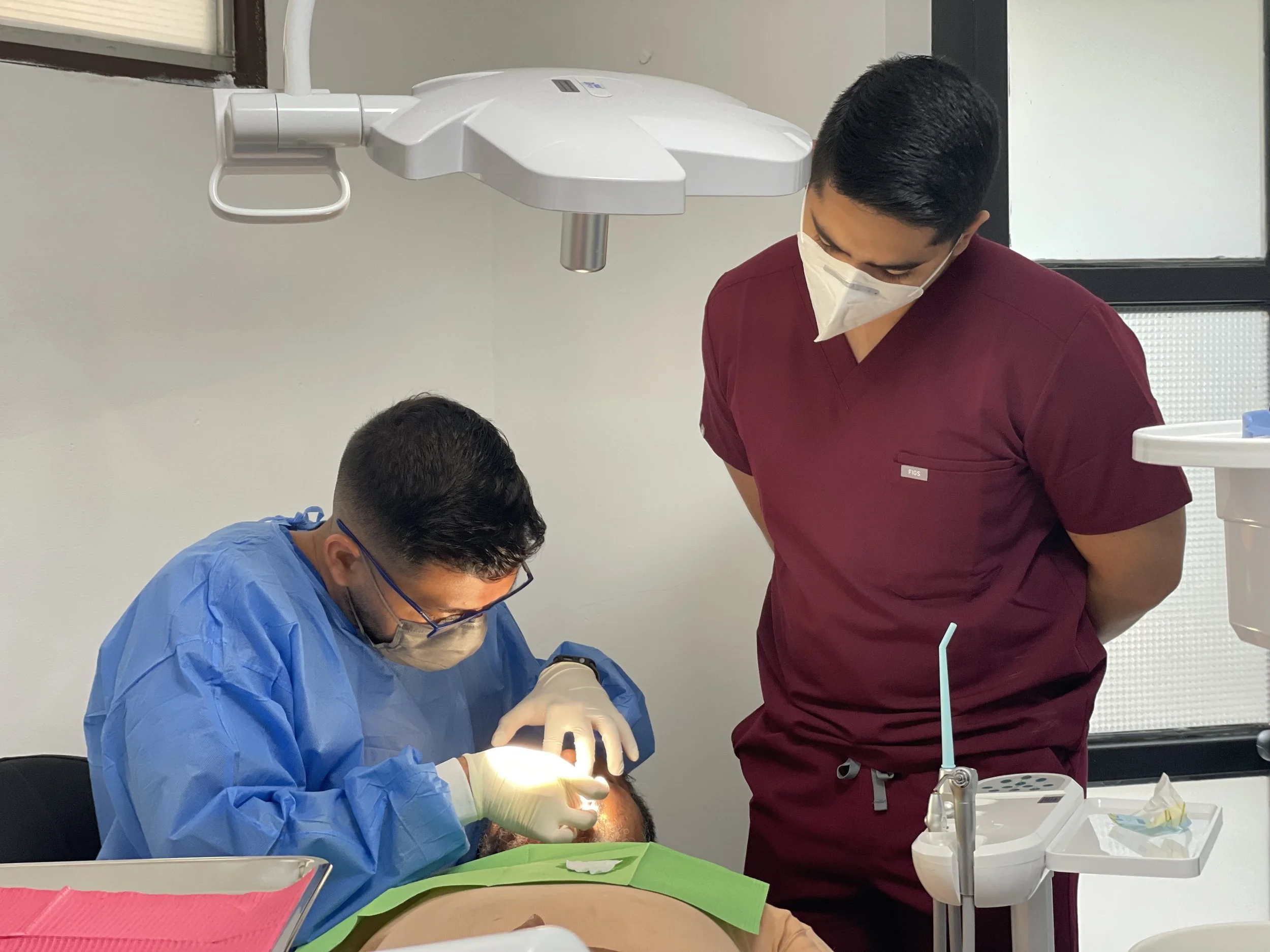 Dentist in blue scrubs and mask performing a dental procedure on a patient lying on the dental chair while another person in maroon scrubs and mask observes in a dental clinic.