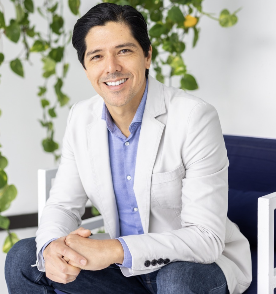 A smiling man with dark hair, wearing a light gray blazer and blue shirt, sitting with hands clasped, in front of green leafy plants.