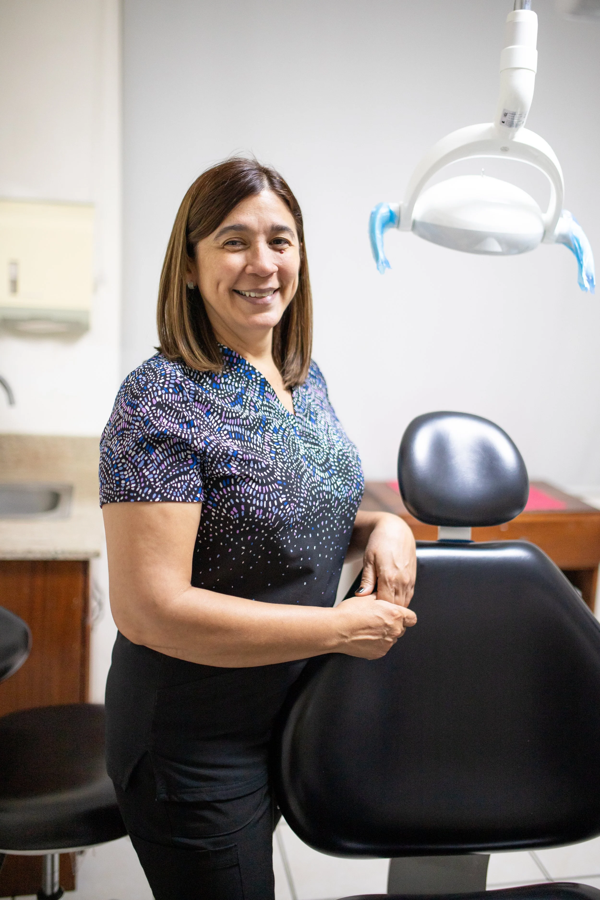 A woman with medium-length brown hair smiling, standing next to a dental chair in a medical office.