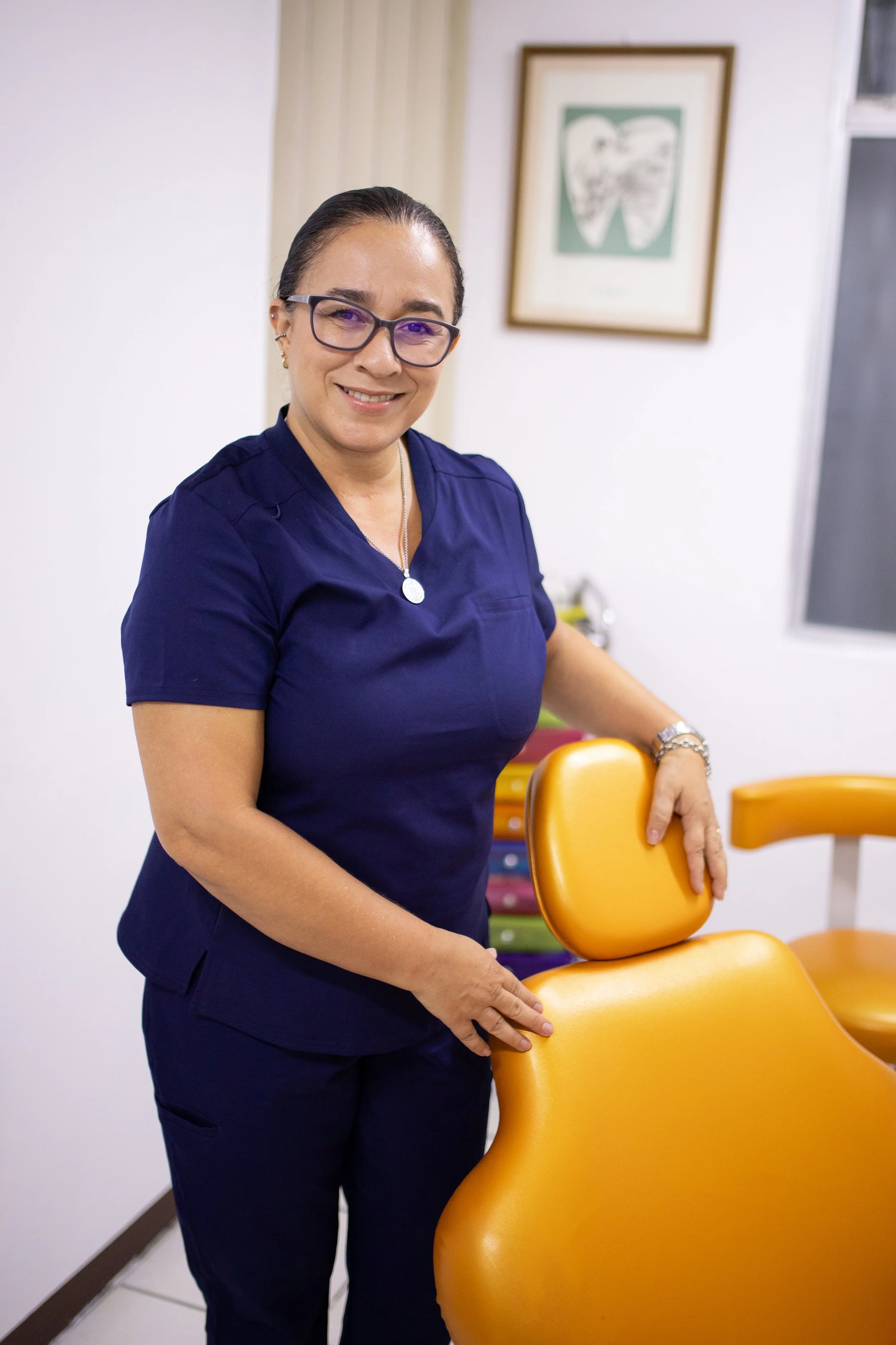 Smiling woman in black medical uniform standing in a dental office with dental chair and equipment in the background.