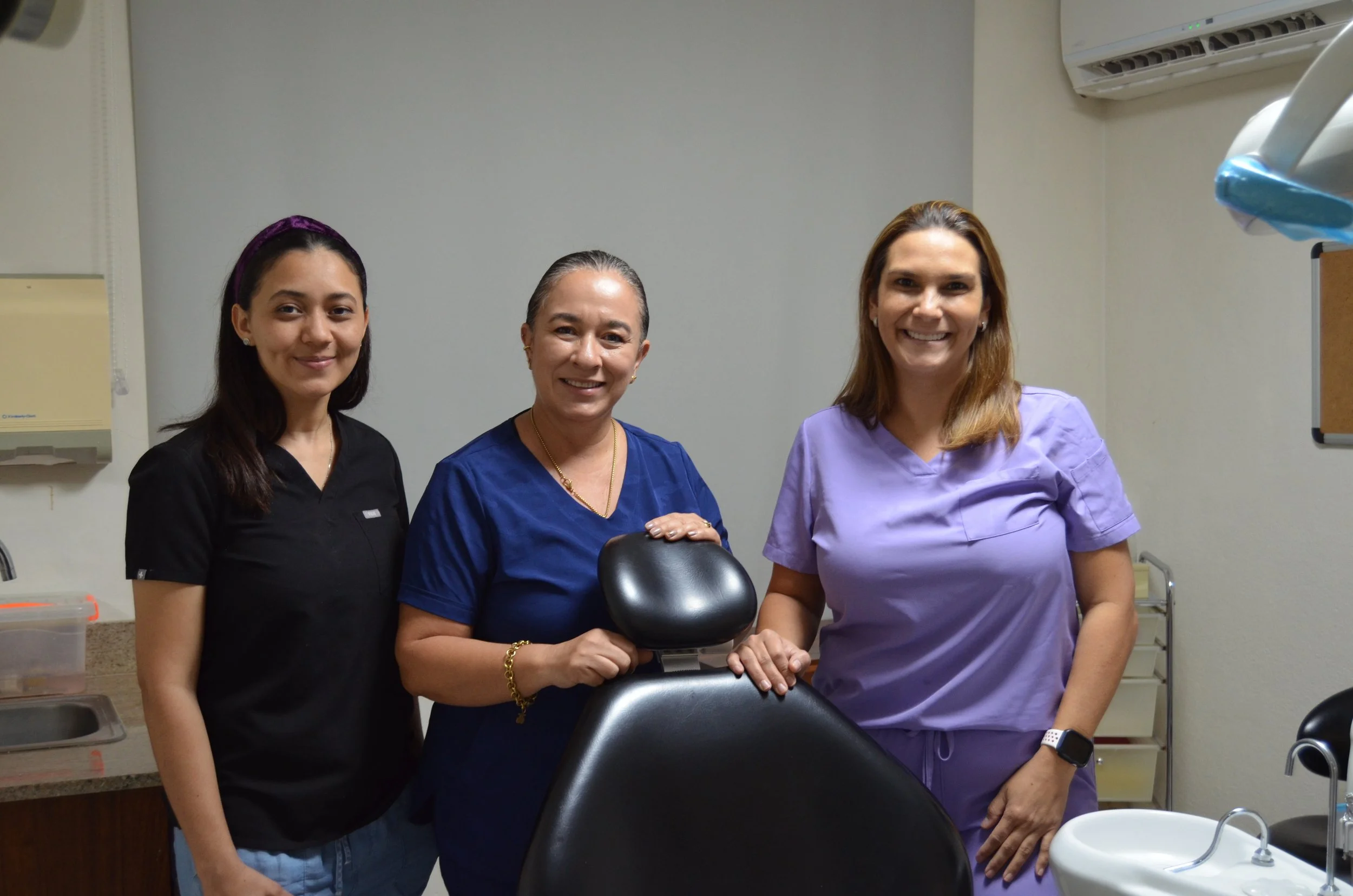 Three women in medical scrubs standing in a hospital or clinic room, smiling at the camera.