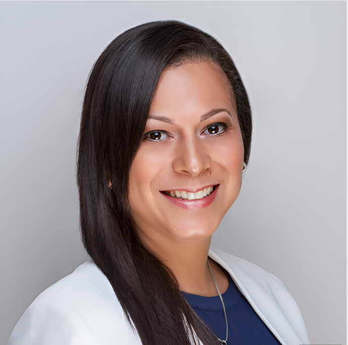A woman with long dark hair smiling, wearing a white blazer and a blue top, against a plain light gray background.