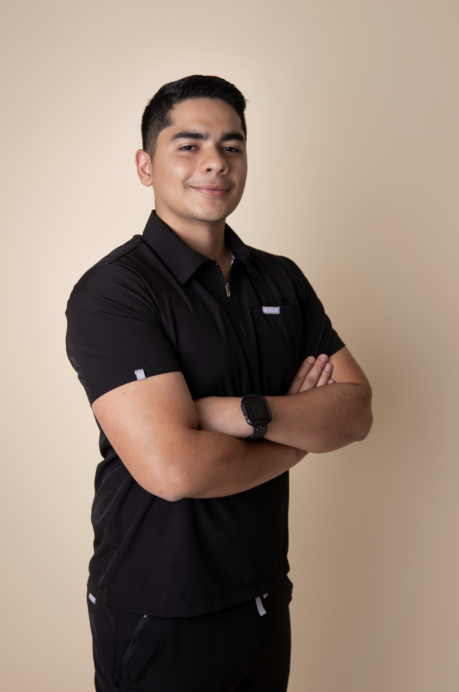 Young man with short dark hair, wearing a black polo shirt and a smartwatch, standing with arms crossed, smiling slightly, against a beige background.