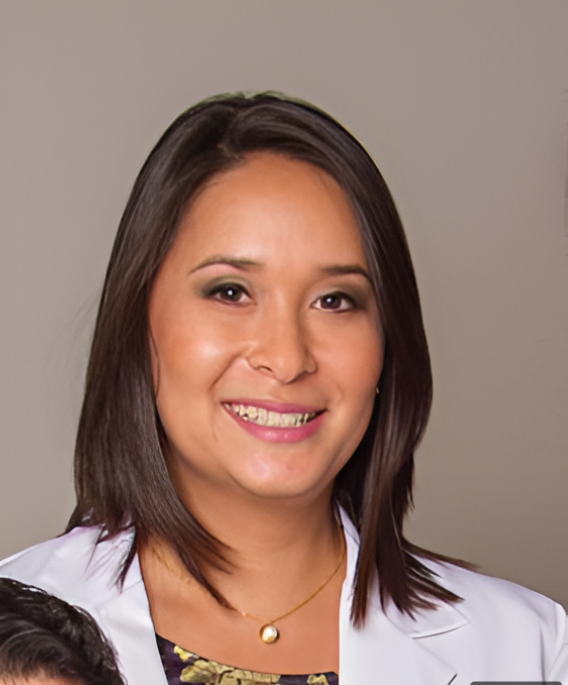 A smiling woman with shoulder-length dark hair, wearing a white lab coat and a necklace, posed against a neutral gray background.