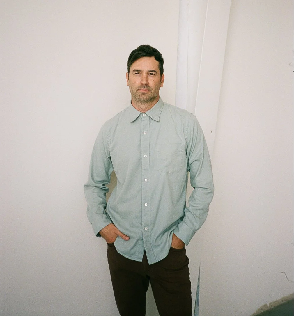 A man with dark hair and a beard stands against a plain white wall, wearing a light gray button-up shirt and dark pants, with one hand in his pocket.