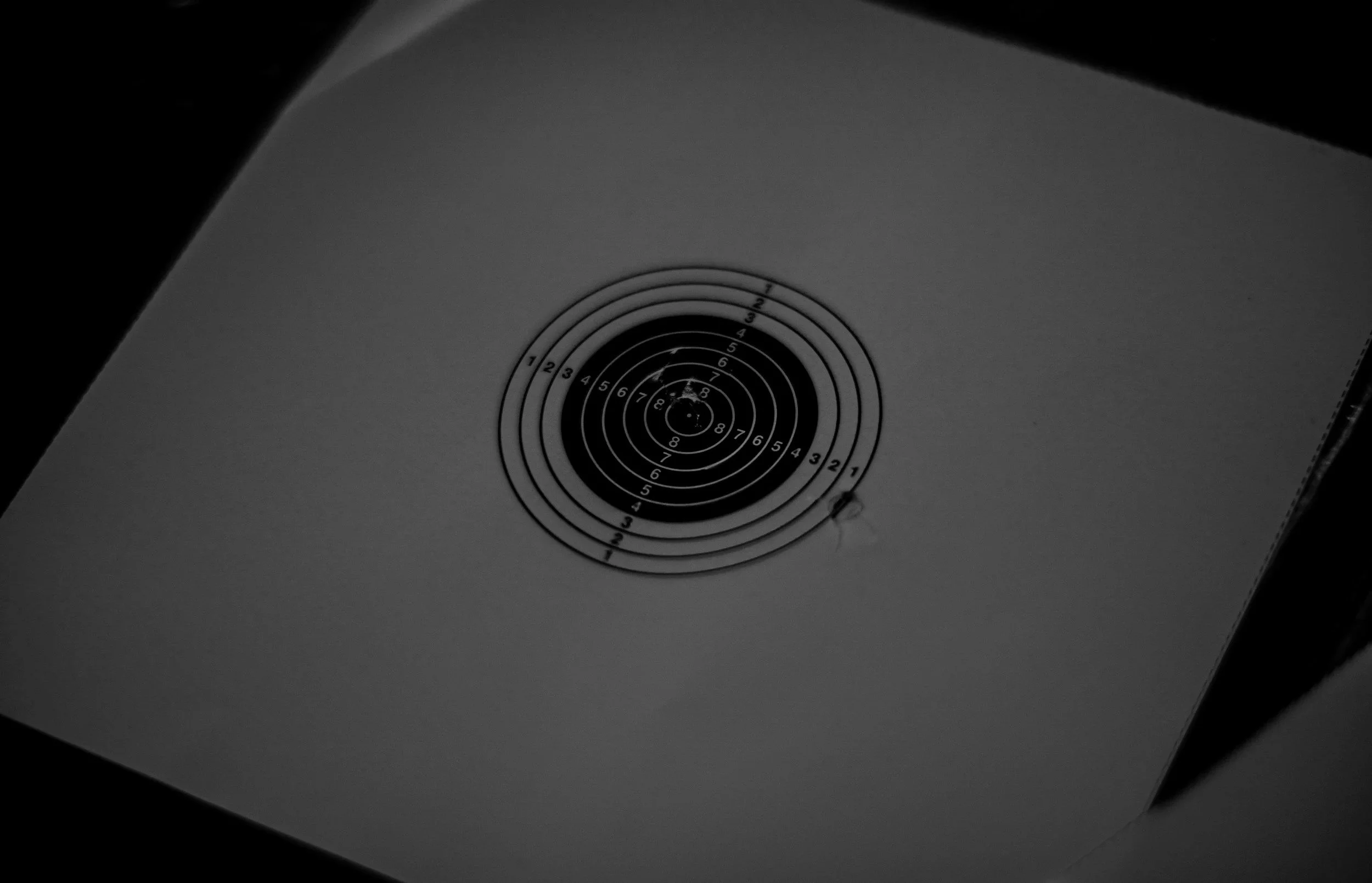 A shooting target with concentric circles numbered from 1 to 10, mounted on a white paper. The target has a few bullet holes, with the most damage near the center.