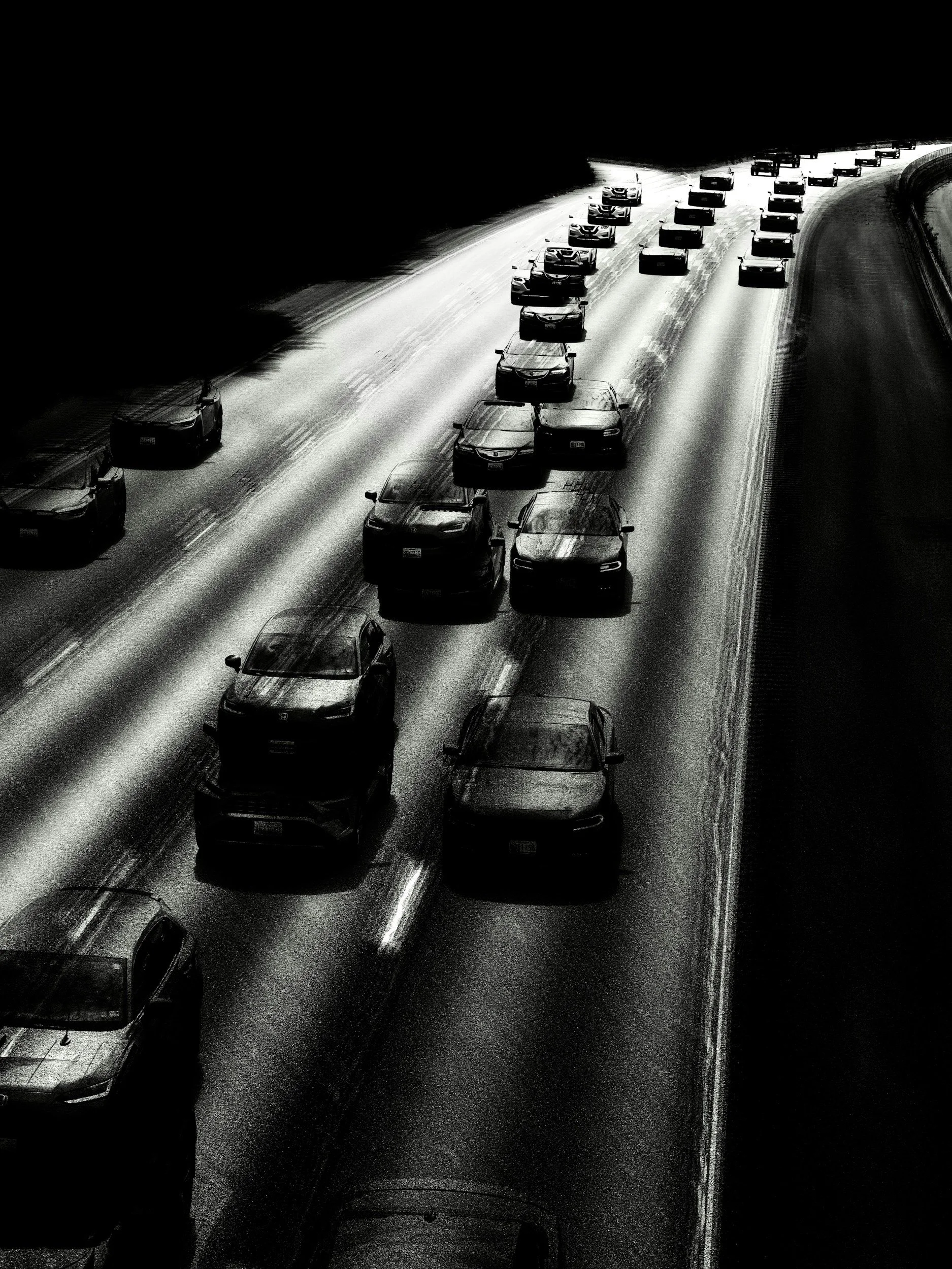 Black and white photo of multiple cars driving on a curved snow-covered highway, some cars appearing blurred due to motion.