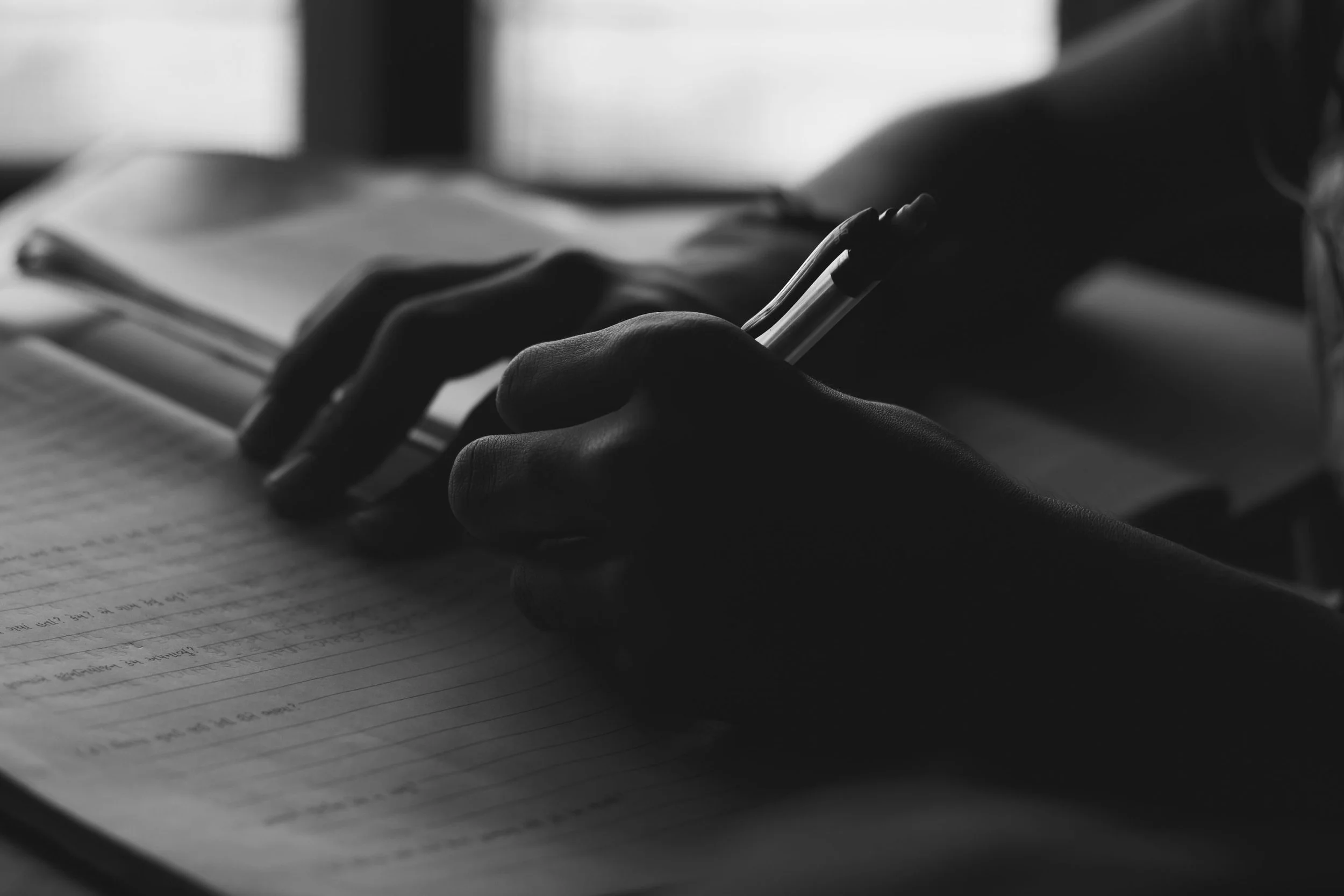 A person's hand holds a pen and writes in a notebook, with notebooks and papers nearby, in black and white.