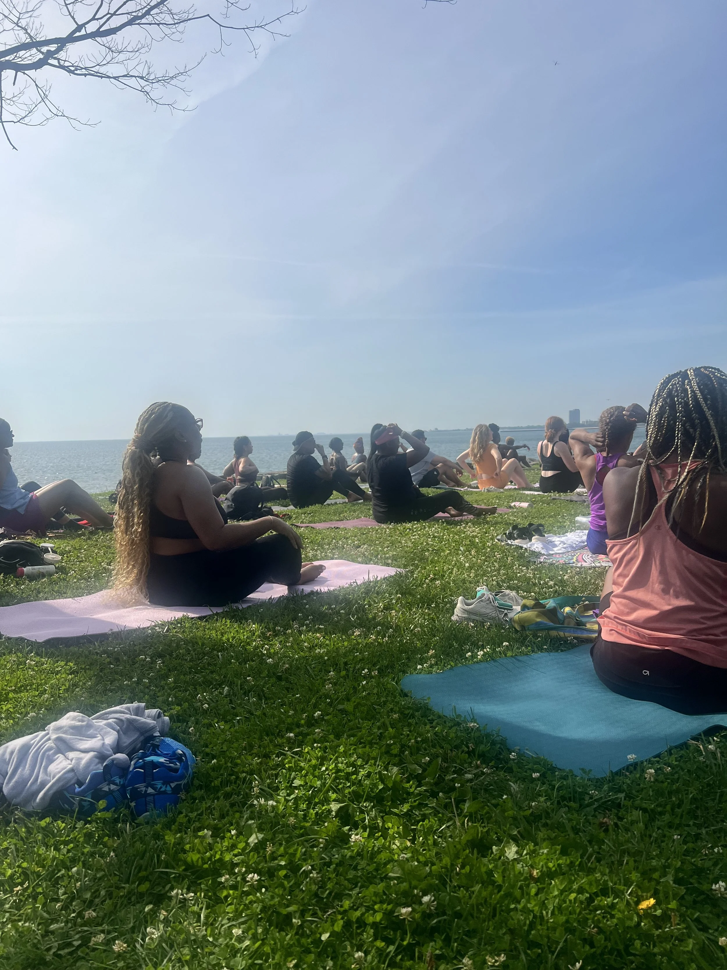 People participating in an outdoor yoga session on a grassy area near the water on a sunny day.