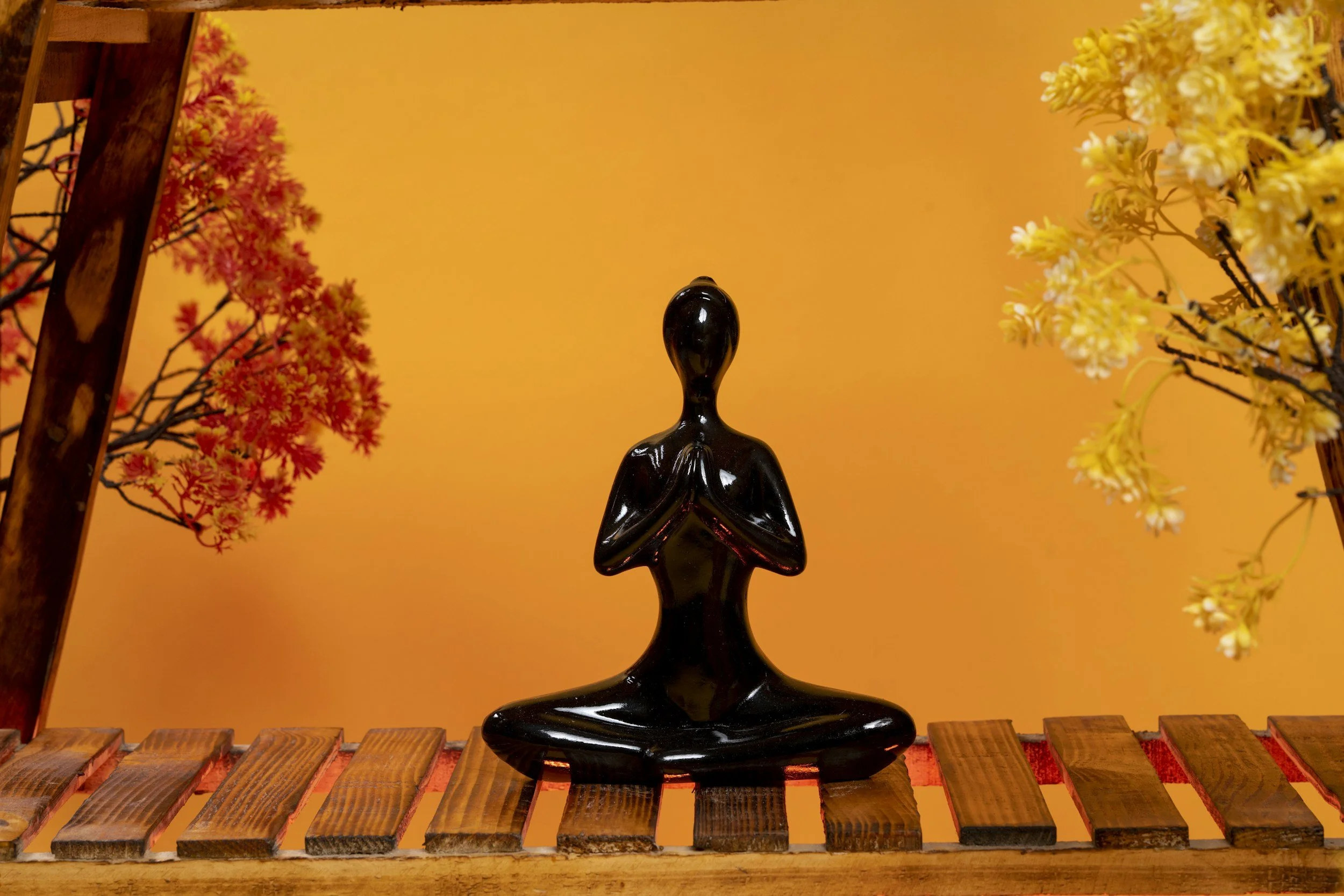 Black sculpture of a person in a meditative pose, sitting cross-legged with hands in prayer position, placed on a wooden surface with colorful artificial flowers and a warm orange background.