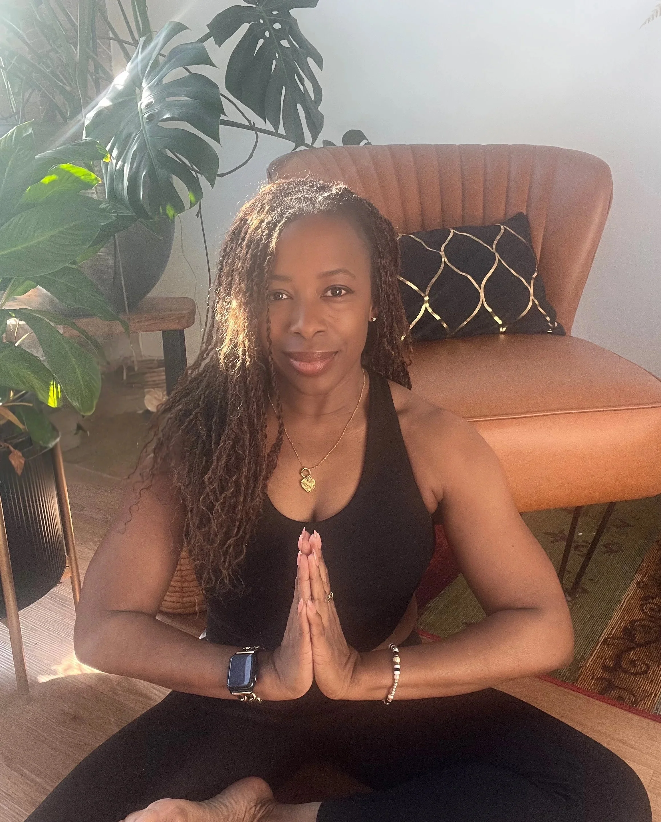 Woman in black tank top practicing yoga in a seated meditation pose with palms together, in a living room with houseplants and a tan armchair behind her.