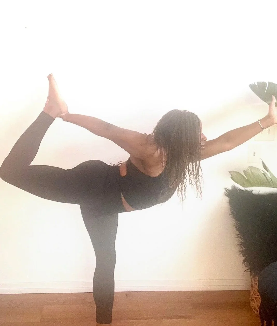 A woman practicing yoga indoors, balancing on one leg while holding the other leg behind her back with her arm extended forward.
